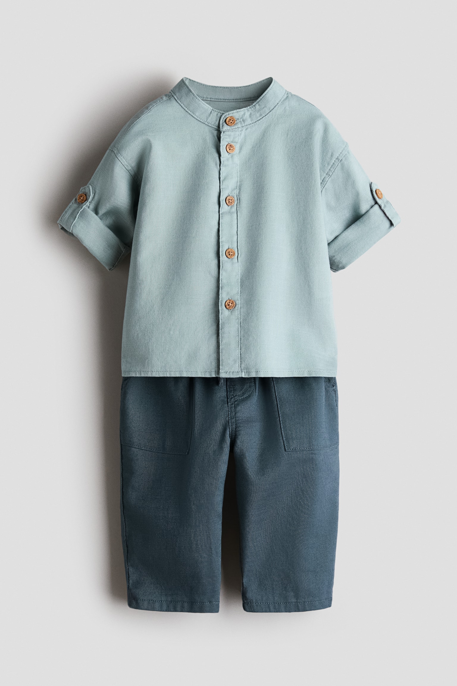 A children's two-piece clothing set featuring a front view of a light sage-green shirt paired with dark teal trousers. The short-sleeved shirt features a band collar, a button-down front with brown buttons, and rolled-up cuffs with button tabs, while the coordinating straight-leg trousers feature front patch pockets.