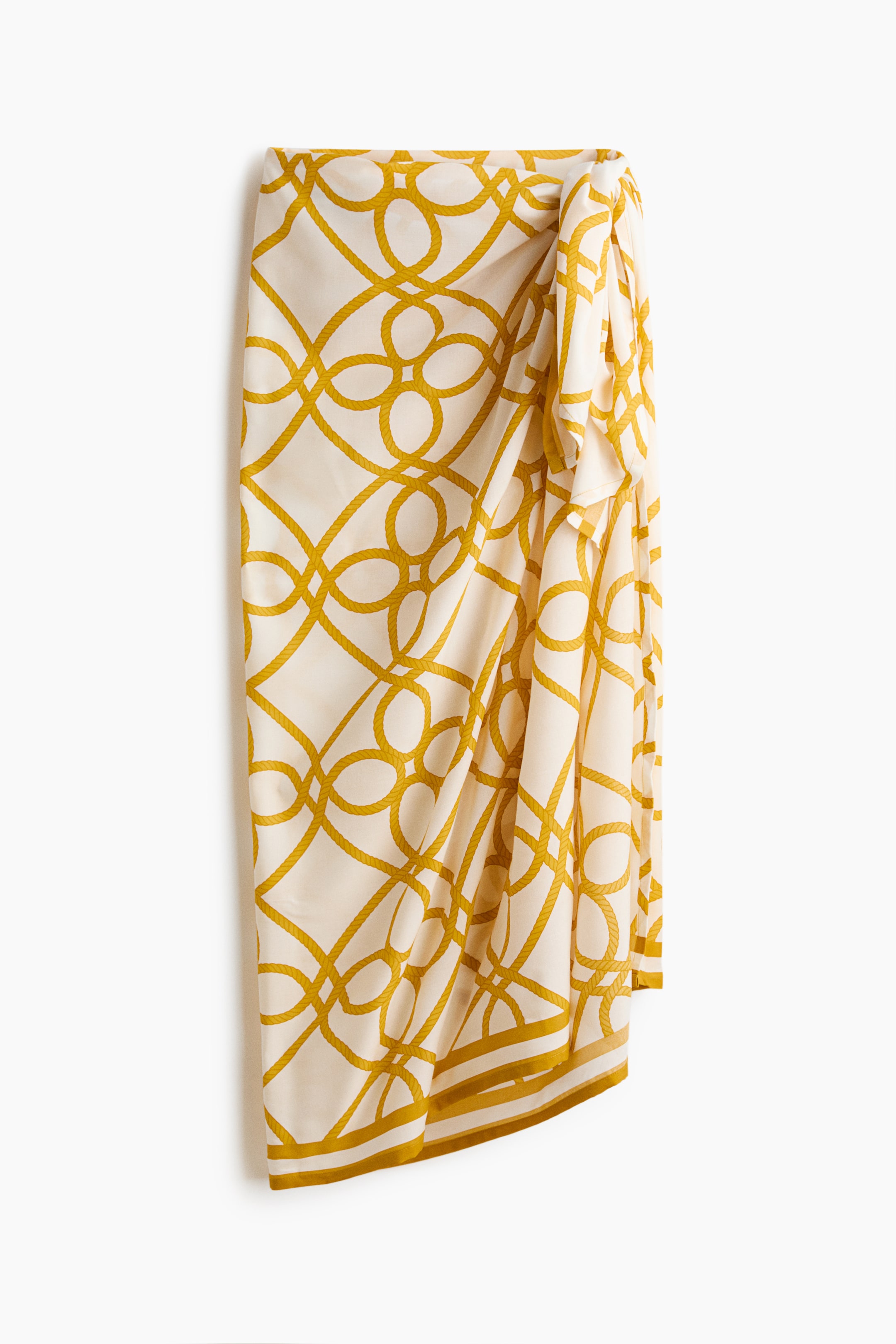 View larger image: A sarong featuring an off-white base with a repeating gold geometric rope pattern. It drapes vertically, showing a tie at the top right, and is finished with a multi-striped border at the bottom.