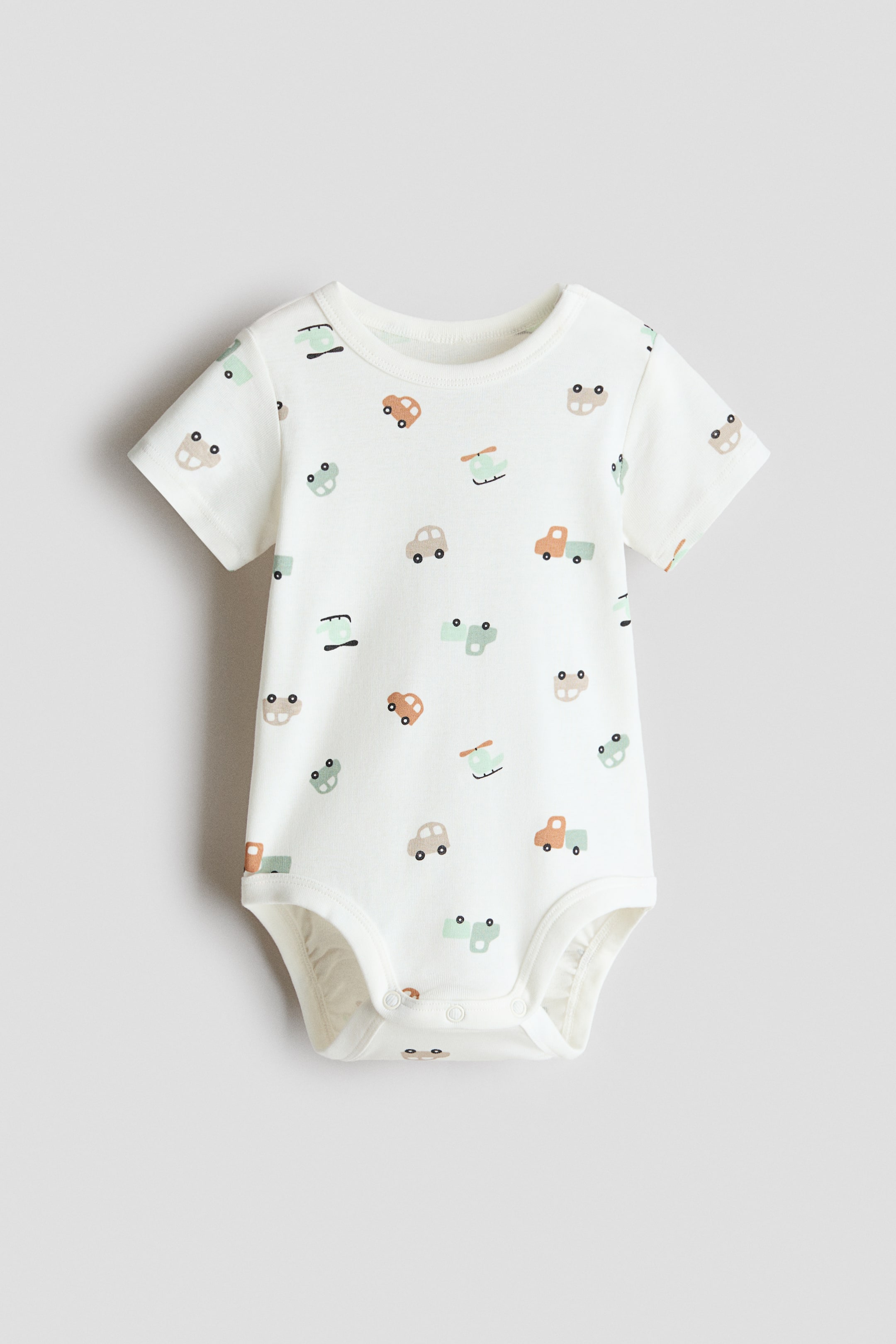 Grotere afbeelding bekijken: Een bodysuit met korte mouwen toont zijn voorkant, voorzien van een all-over print van gestileerde voertuigen in gedempte bruine, oranje en groene tinten op een crèmekleurige achtergrond. Het is ontworpen met een afgewerkte ronde halslijn, en drukknopen sluiten op één schouder en bij het kruis.