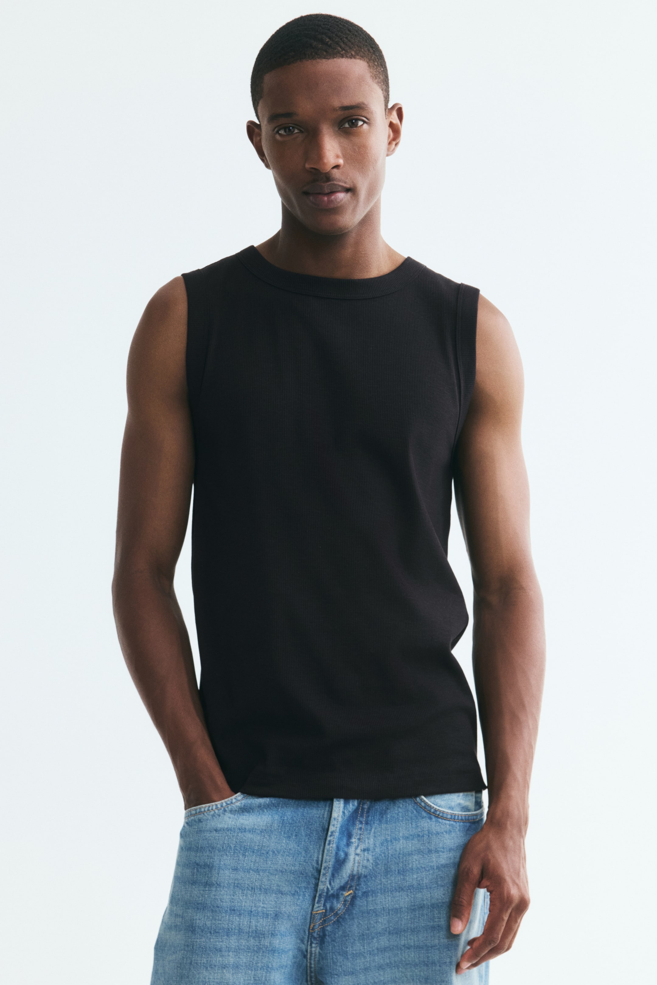 View larger image: A black ribbed-knit vest top featuring a round neckline and a slim-fit silhouette is worn by the model. The top is styled with light blue jeans, and the model has one hand in a pocket.