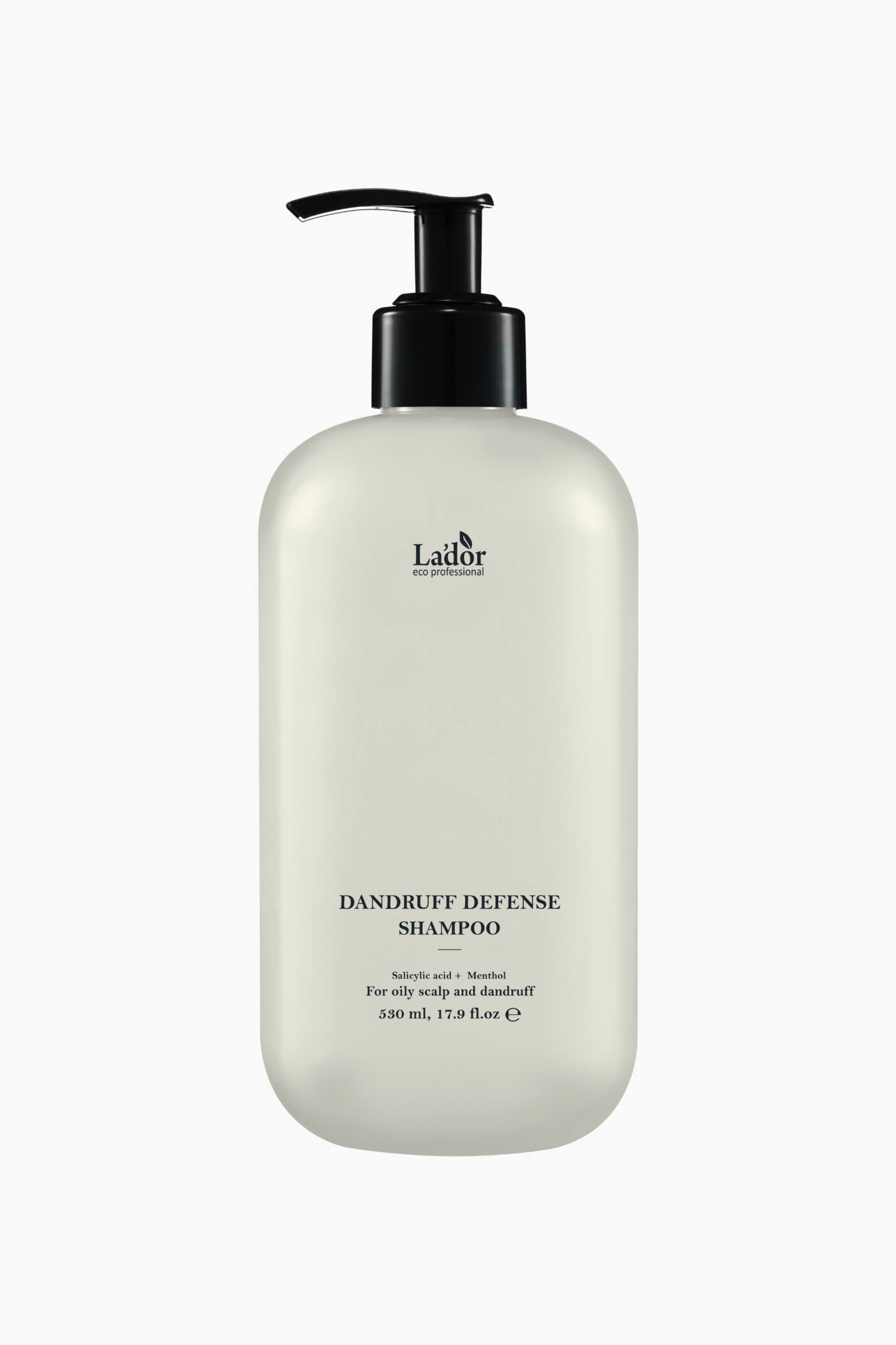 Se større billede: En off-white, rektangulær shampoo flaske med afrundede kanter står oprejst og har en sort pumpe dispenser. Forsiden af flasken viser "L'ador eco professional" logoet, "DANDRUFF DEFENSE SHAMPOO", samt tekst der beskriver dens ingredienser og volumen.