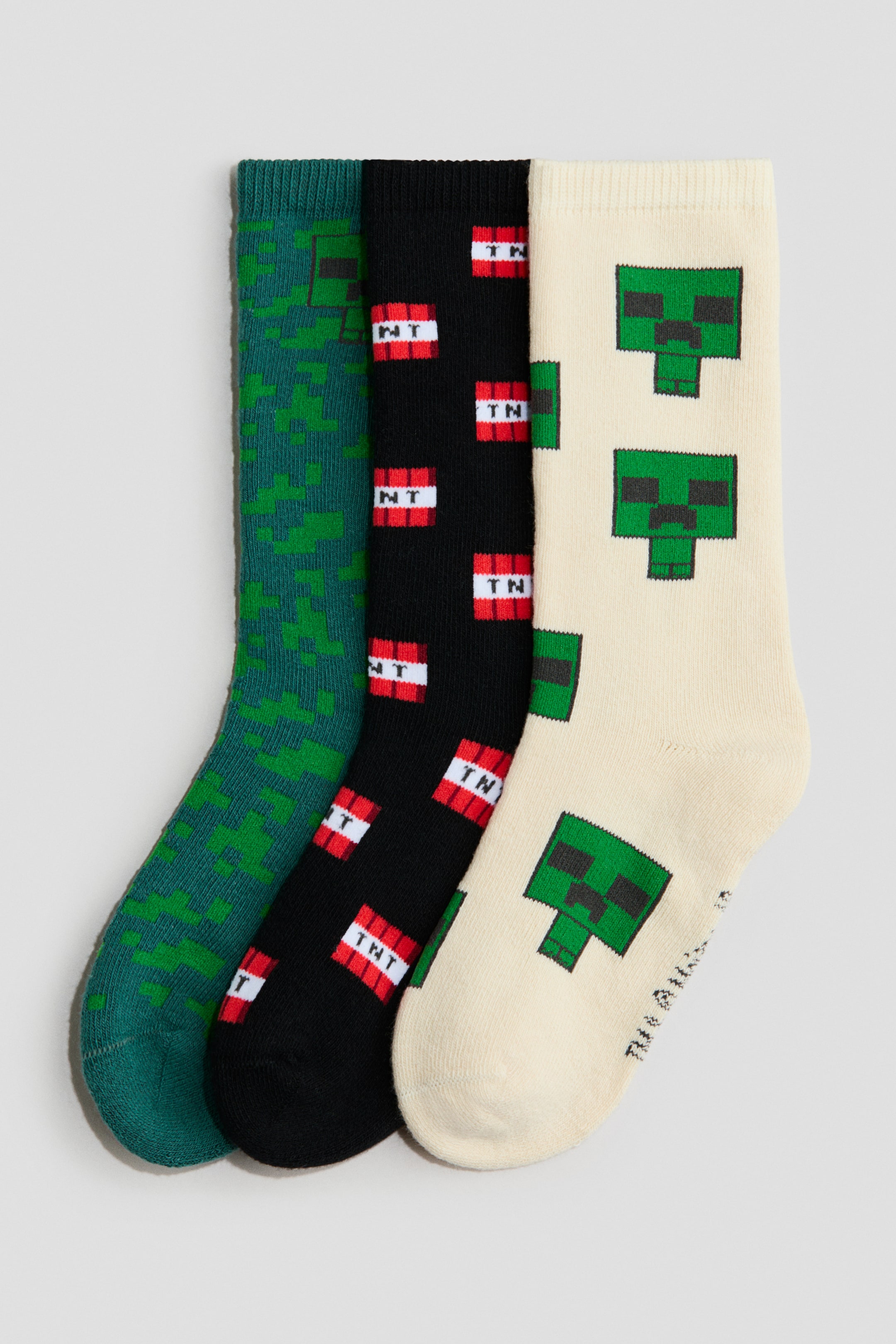 View larger image: A three-pack of knit crew socks is displayed flat, featuring ribbed cuffs and pixelated intarsia designs. The set includes a green sock with a pixelated tonal pattern, a black sock with red and white "TNT" block motifs, and an off-white sock featuring green pixelated face designs.