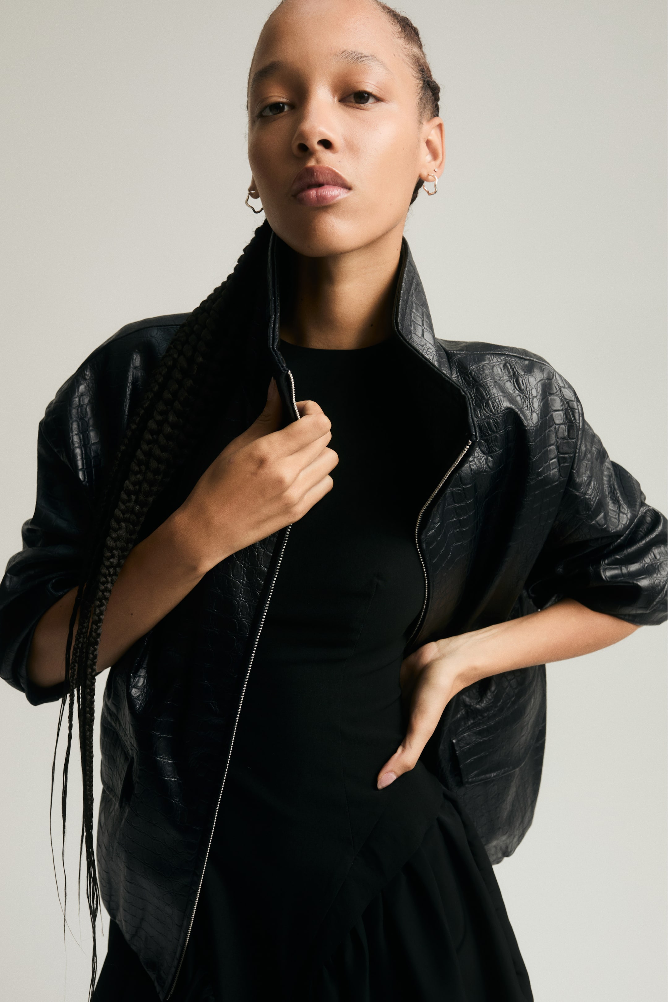 Größeres Bild anzeigen: Eine oversized schwarze Jacke aus beschichtetem Stoff mit Krokodilstruktur wird vom Model über einem schwarzen Oberteil getragen. Das Model hält den Rand des Stehkragens mit einer Hand, während die andere Hand auf der Hüfte ruht, wodurch die überschnittenen Schultern und der Frontreißverschluss der Jacke betont werden.