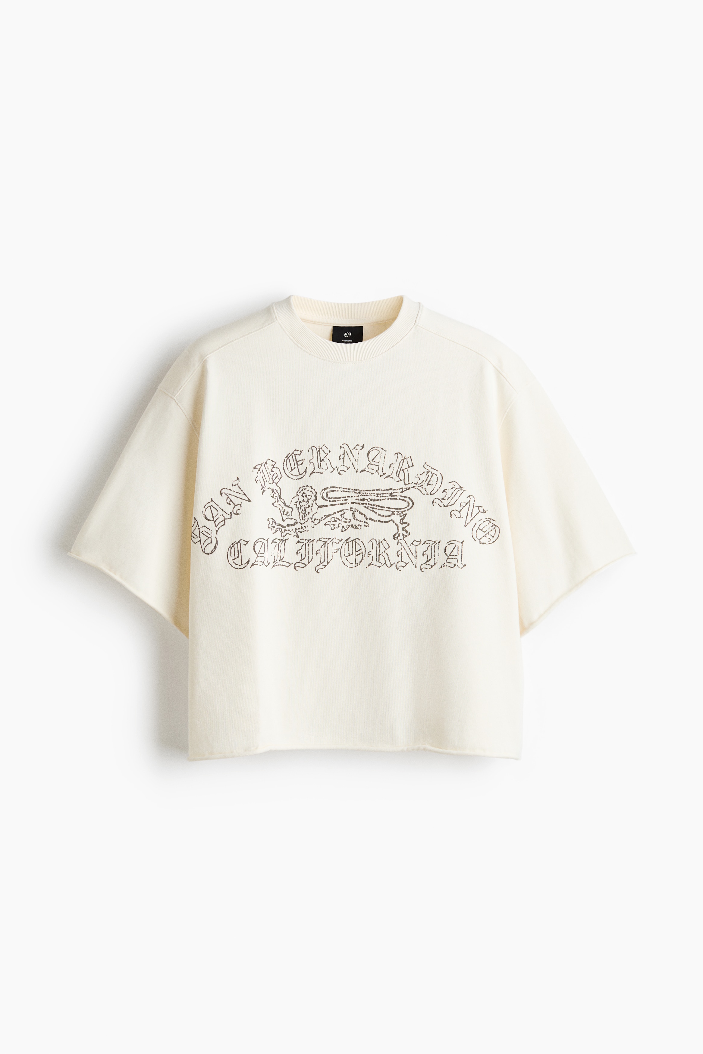 An off-white, boxy-style T-shirt featuring a printed graphic on the front is being worn by the model. The oversized tee is designed with dropped shoulders, a round ribbed neckline, and raw, rolled edges at the cuffs and hem, styled with black jeans and a baseball cap.