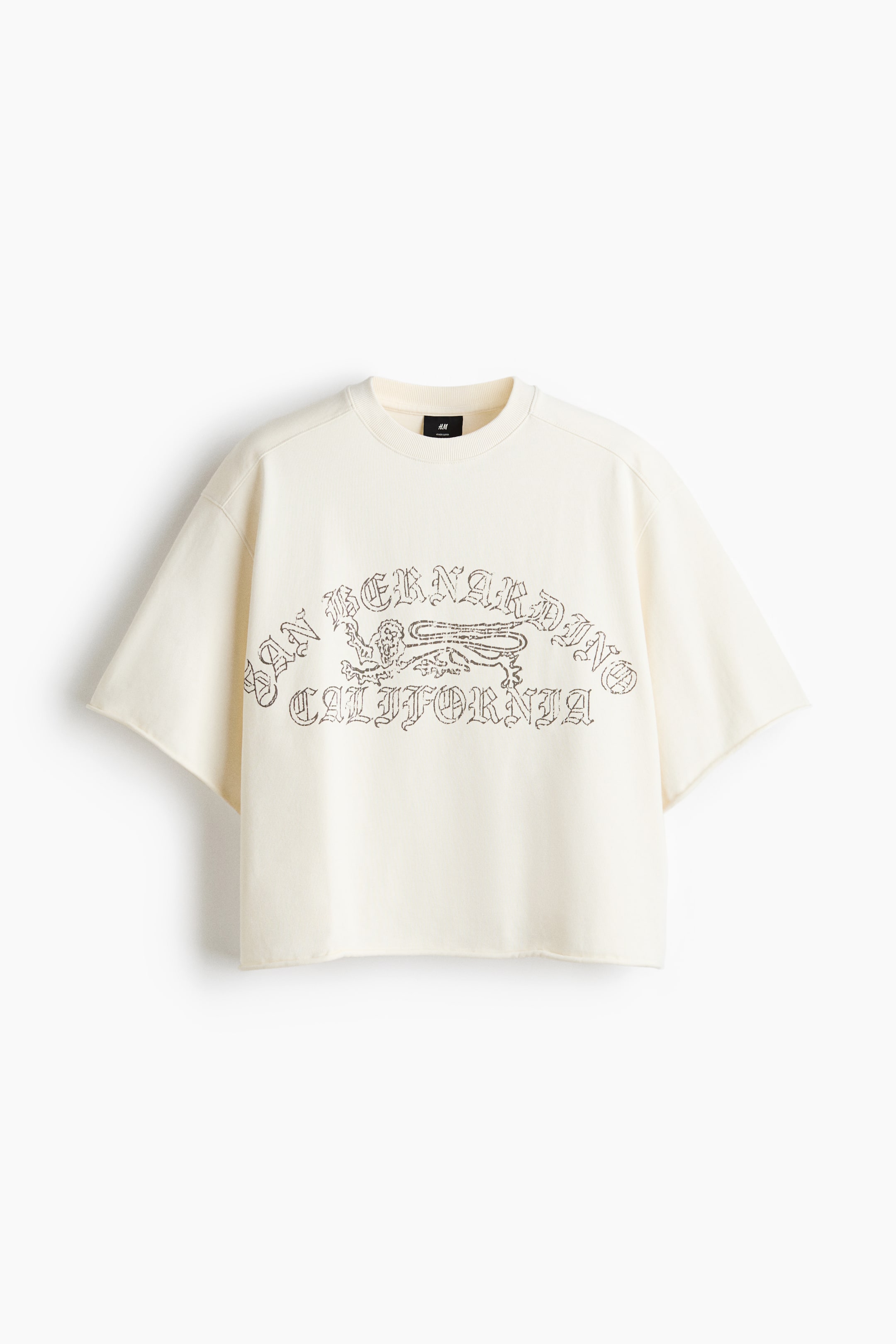 View larger image: The cream short-sleeved sweatshirt features a ribbed crew neck, exposed shoulder seams, and wide, raw-cut sleeve edges. An arched graphic print reading 'SAN BERNARDINO CALIFORNIA' with a mythical creature is visible on the front, above a raw-cut cropped hem.