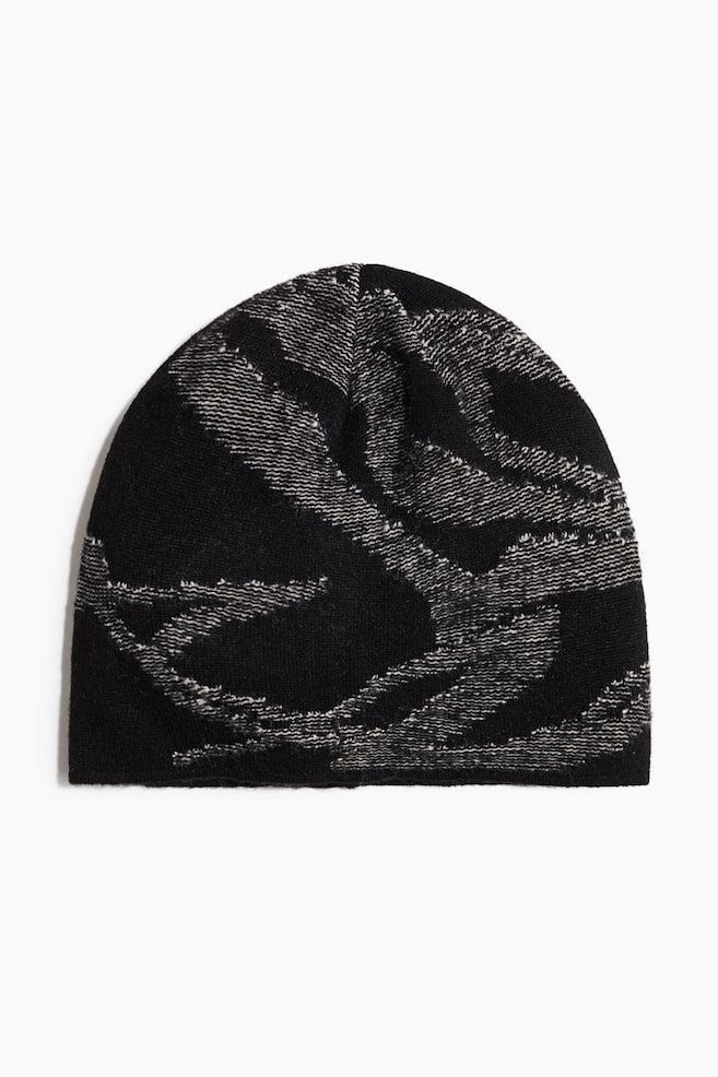Eine StrickmÃ¼tze ist von vorn zu sehen und zeigt vor einem einfarbig schwarzen Hintergrund ein abstraktes, wellenfÃ¶rmiges Muster in einem kontrastierenden grauen und weiÃen Marl-Strick. Die MÃ¼tze verfÃ¼gt Ã¼ber eine abgerundete Krone und einen sauberen, umgeschlagenen unteren Saum ohne Umschlag. - 1