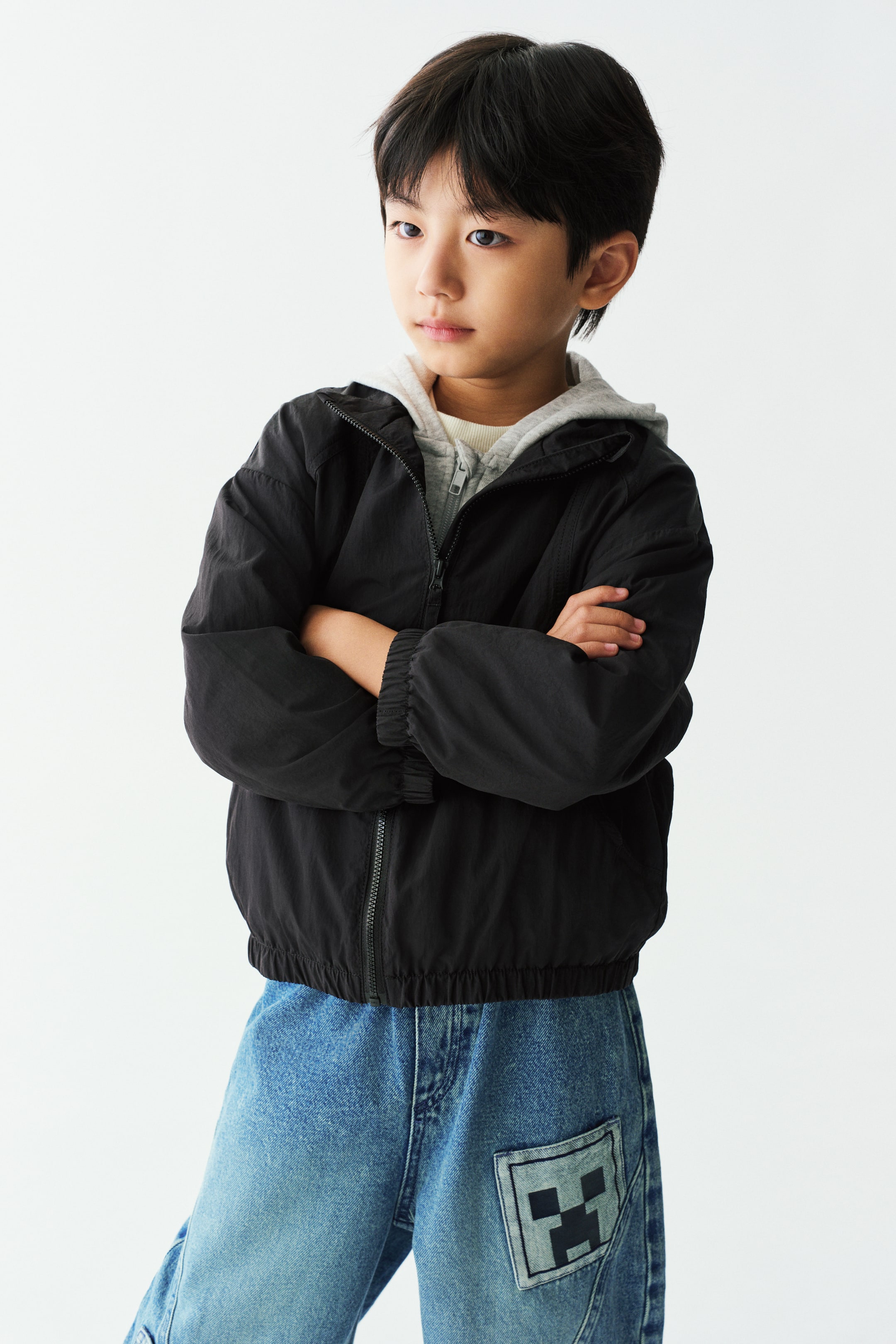 View larger image: A black nylon windbreaker with a front zip, dropped shoulders, and elasticated cuffs and hem is worn by the model with arms crossed. The loose-fit jacket is layered over a light grey zip-up hoodie and styled with blue jeans.