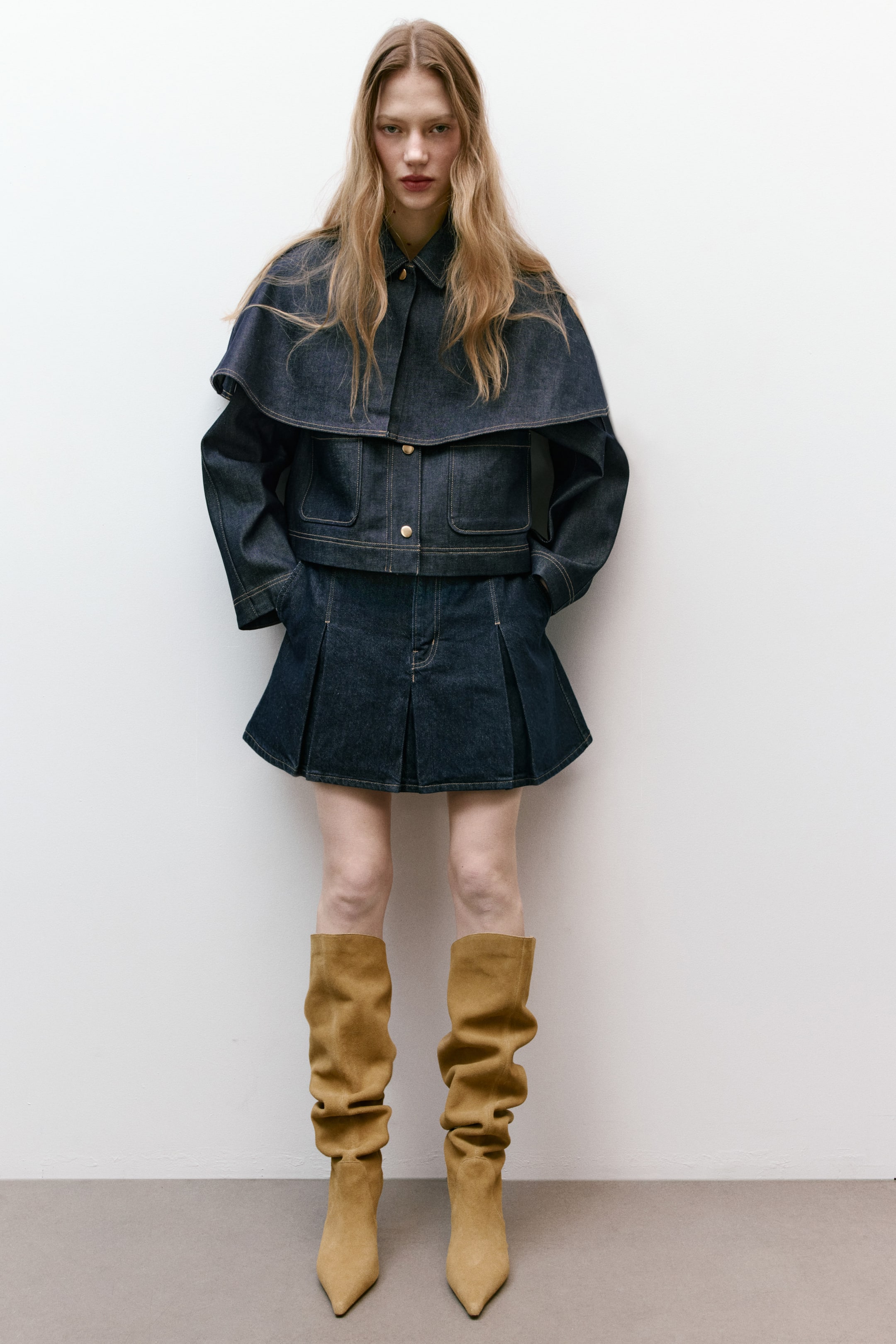 View larger image: Presenting a dark wash denim jacket paired with a matching pleated mini skirt. The cropped jacket showcases a layered cape over the shoulders, a pointed collar, two chest flap pockets with gold-toned buttons, and contrast stitching throughout. The mini skirt features a pleated design.