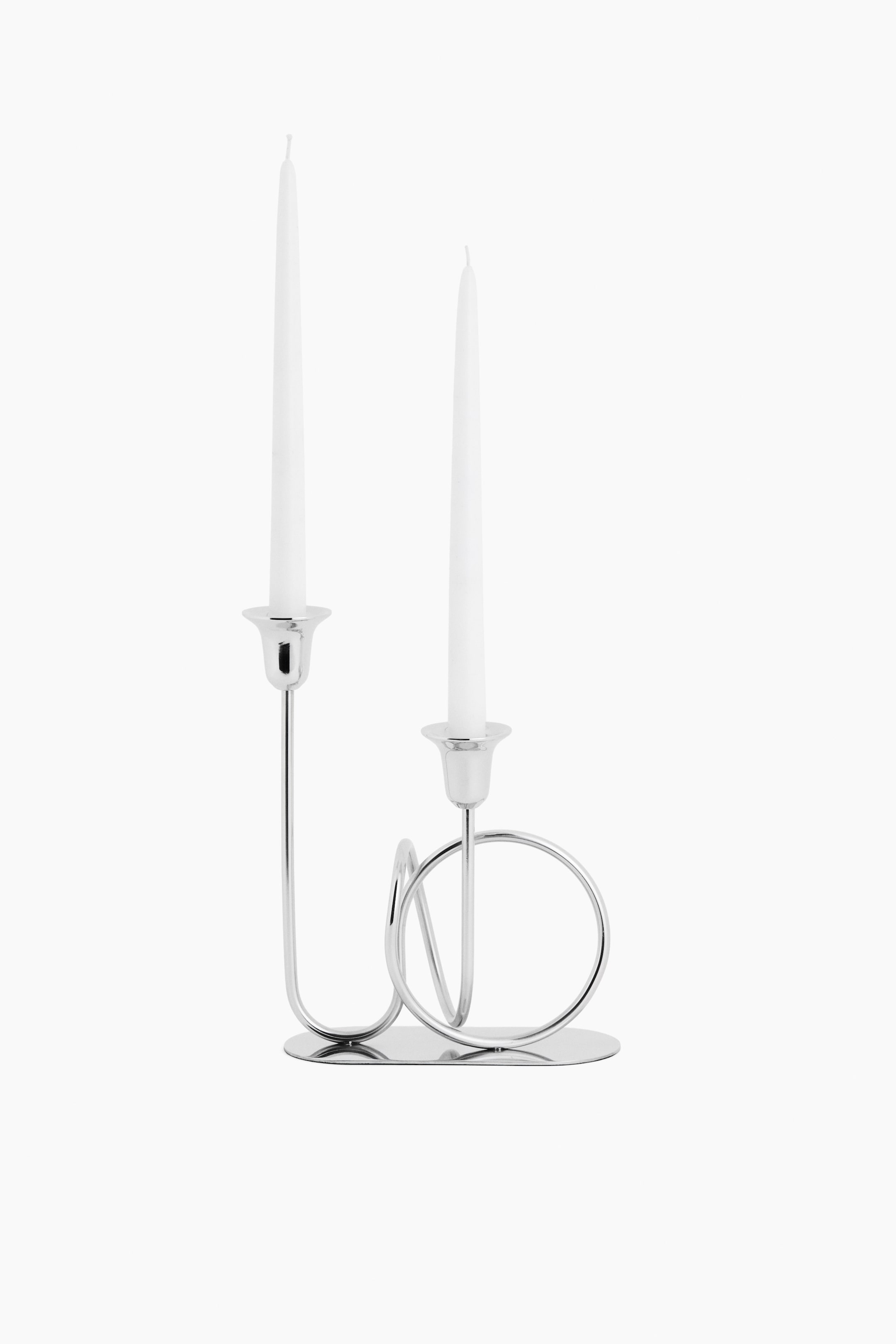 View larger image: A stainless steel candelabra is shown from the front, standing on a flat, oval base, featuring a polished finish with a knot-inspired design of looping metallic rods. It holds two tall, white taper candles in tulip-shaped cups at staggered heights.