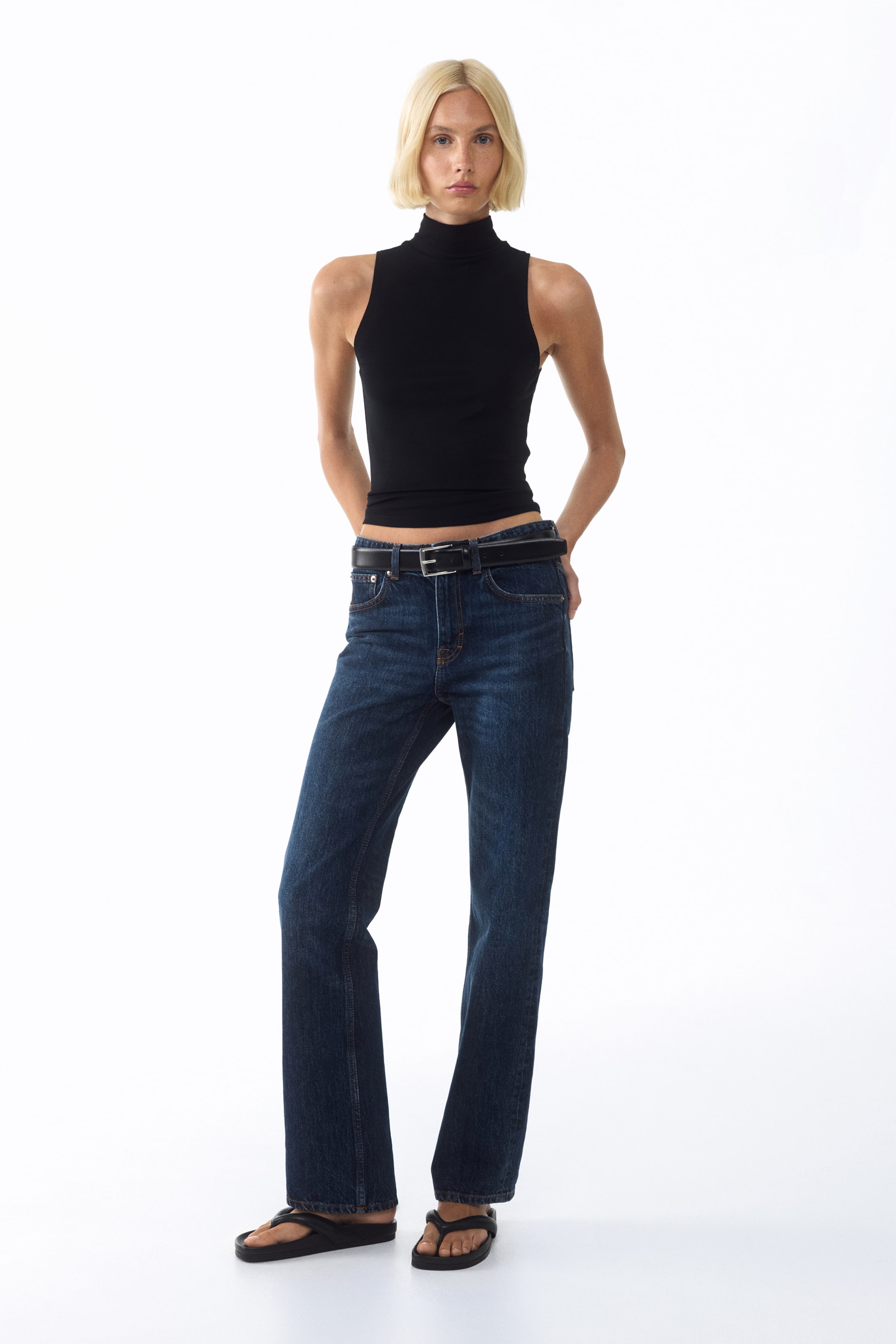 Visualizza immagine più grande: Un top nero senza maniche a collo alto, caratterizzato da una silhouette aderente e rivolto in avanti, è indossato infilato in un paio di jeans a gamba dritta blu scuro. I jeans sono accessoriati con una cintura nera che presenta una fibbia in tonalità argento, e dei sandali infradito neri completano l'outfit.