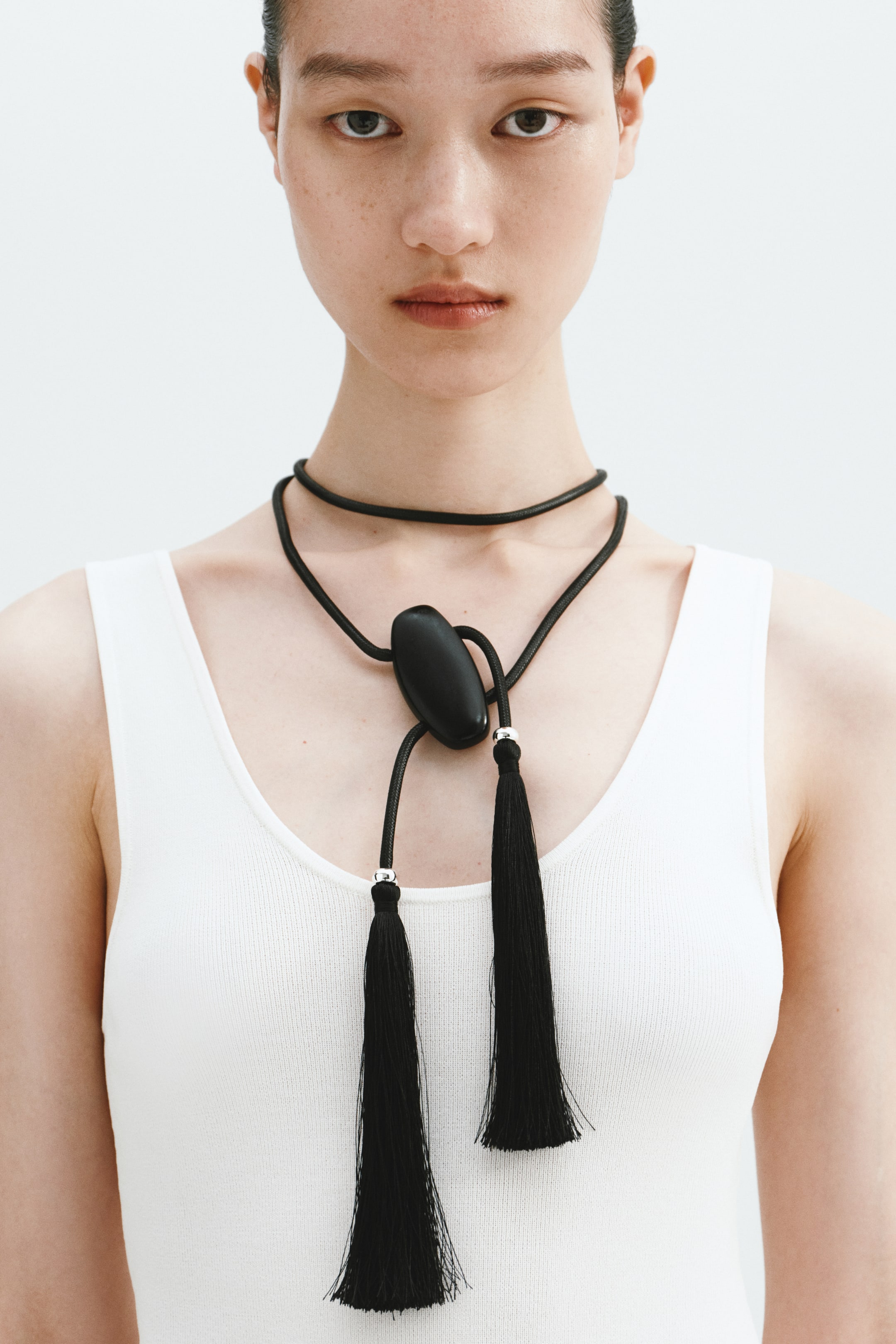 View larger image: The model is wearing a black cord necklace featuring a prominent oval-shaped black sliding bead at the front, complemented by two long black tassels with silver-toned caps at the ends.
