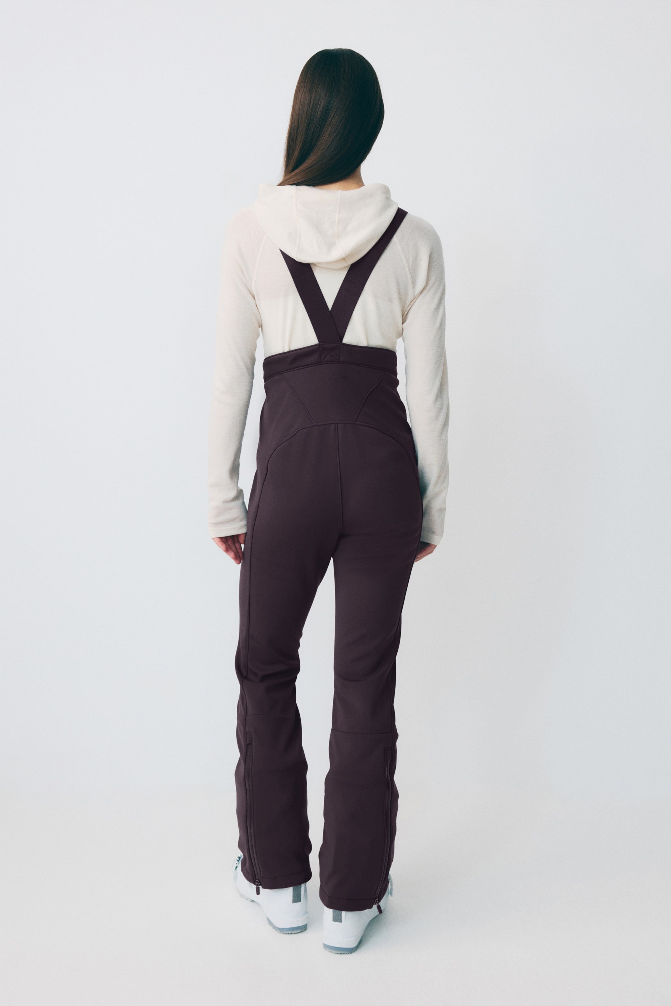 Agrandir l'image: Les pantalons de ski à bretelles couleur prune foncée sont présentés de l'arrière, avec des bretelles en forme de V sur un haut à manches longues à capuche de couleur claire. Les pantalons sont ajustés haut à la taille et comprennent des fermetures éclair aux chevilles.