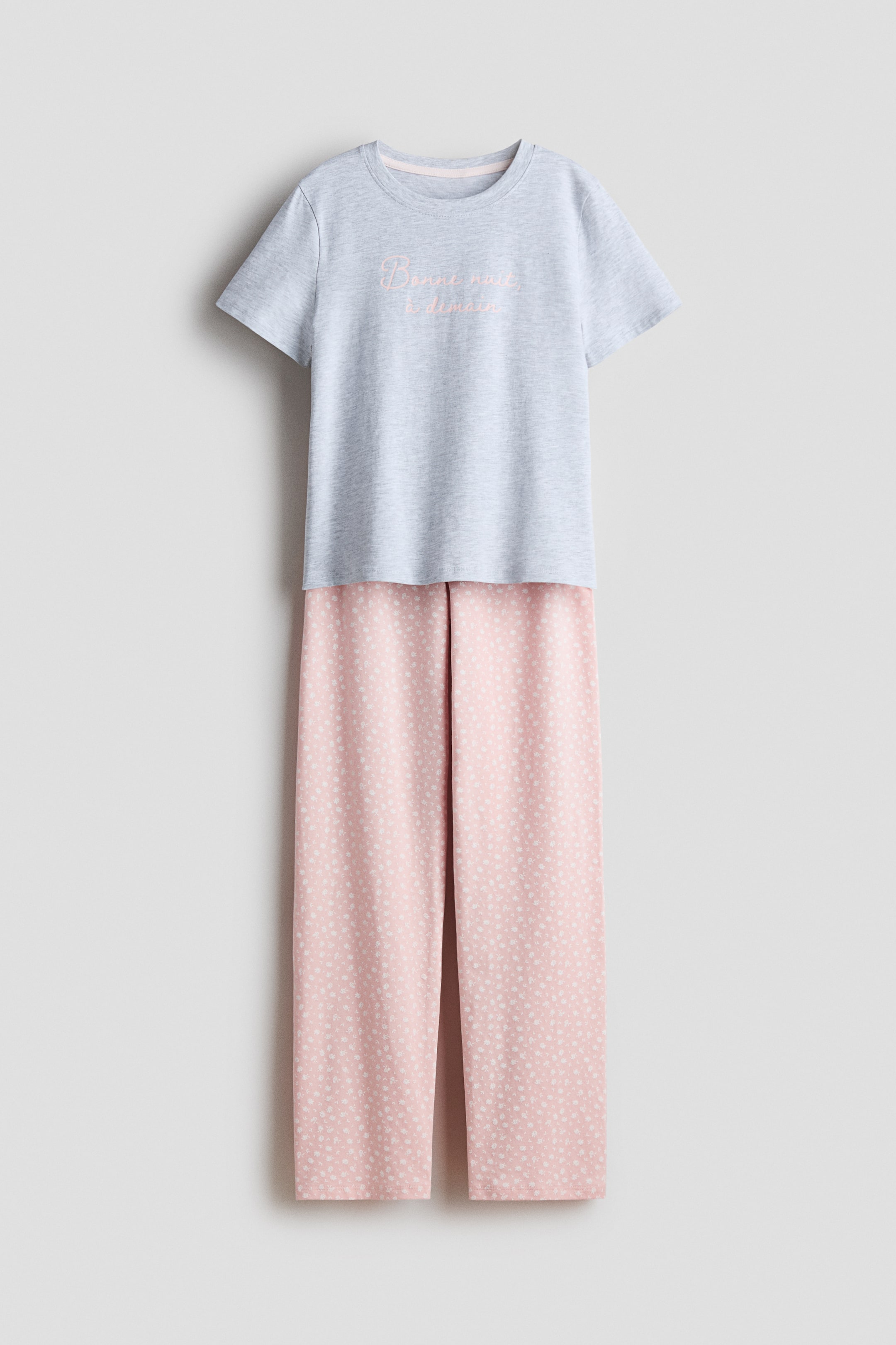 Grotere afbeelding bekijken: Een pyjama set met een heather grijze top met korte mouwen en bijpassende roze bedrukte broek, aan de VOORKANT. De top heeft een ronde, geribde halslijn met roze geborduurde tekst 'Bonne nuit � demain' op de borst, en de broek heeft een klein wit stippenpatroon op roze stof met lange, rechte pijpen.