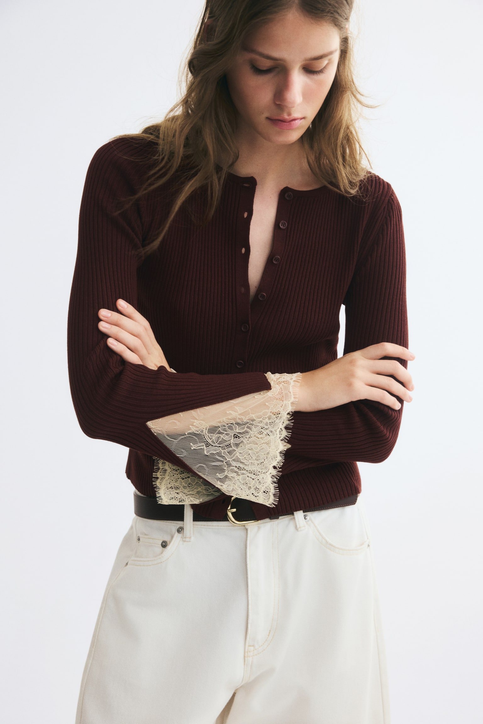 A dark brown ribbed knit cardigan with a button-down front is worn by the model, who is crossing her arms to showcase the flared cuffs trimmed with cream-coloured lace. The cardigan is styled with off-white trousers and a dark belt. - 1