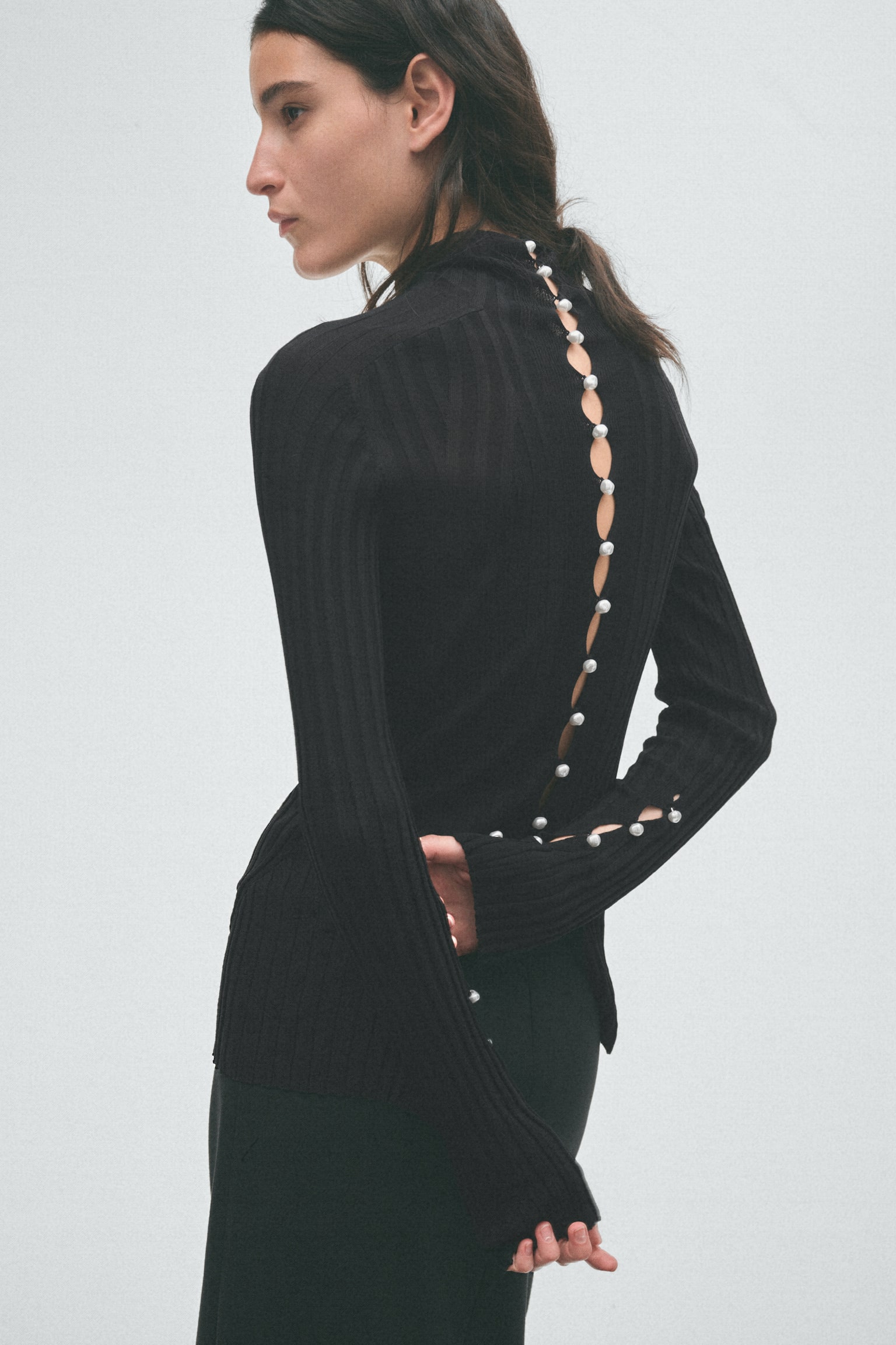 A black ribbed knit top showcases a high neck with three heart-shaped cutouts at the back. The torso is adorned with diagonal ribbing, while its long sleeves boast open cuffs with five small pearl buttons running up each side. - 1