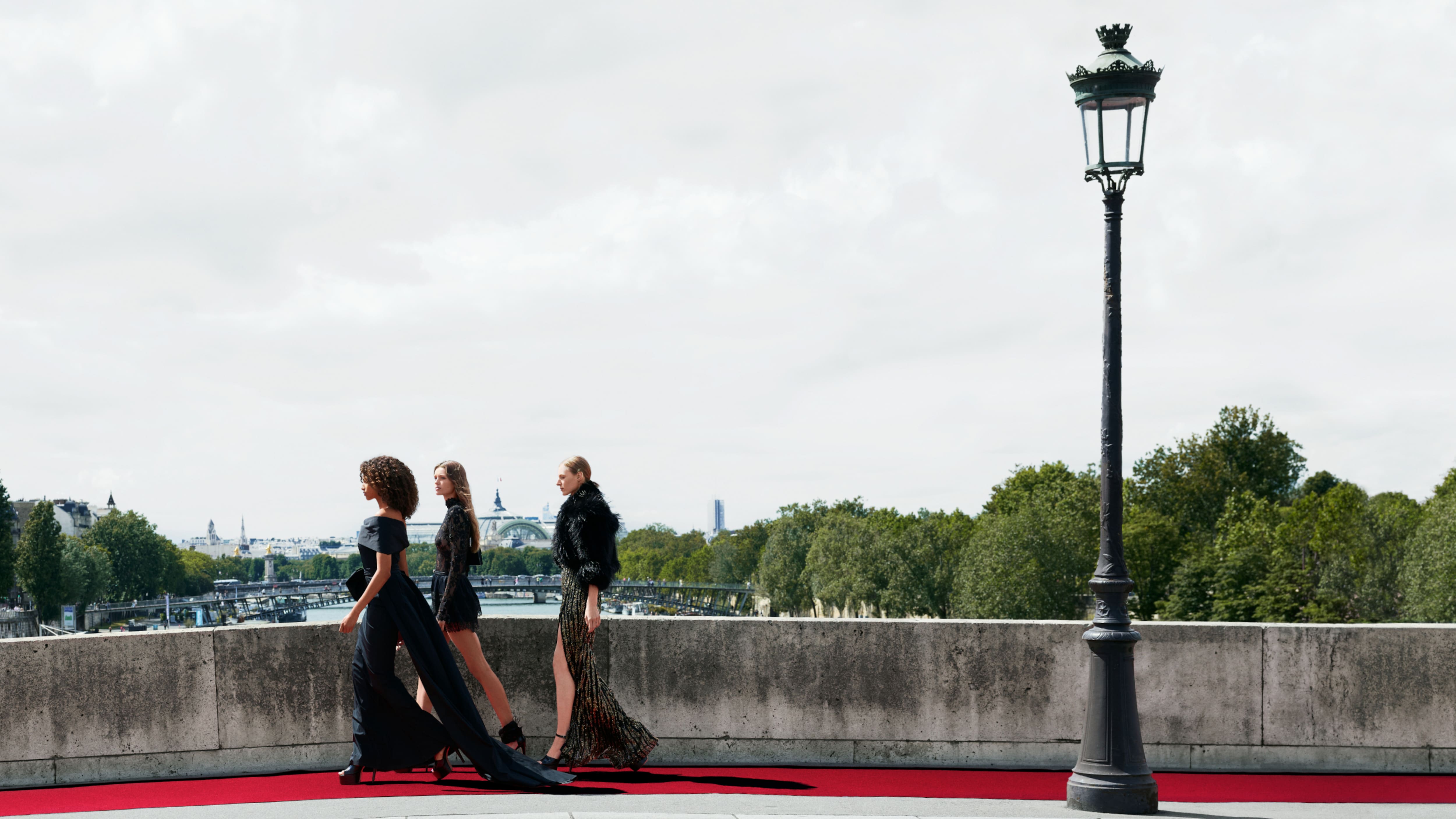 Models in black tailored gowns from the Elie Saab H&M collaboration walking on a red runway on Paris bridge overlooking the Seine.